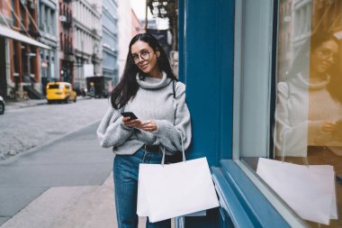 Gözlüklü, kot pantolon ve süveter giyen çekici, narin bir kadın. Şehir caddesinde duvara yaslanmış kameraya bakıyor.