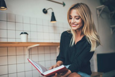 Satisfied blond haired young woman with radiant smile in stylish casual green shit reading book while resting at modern apartment 