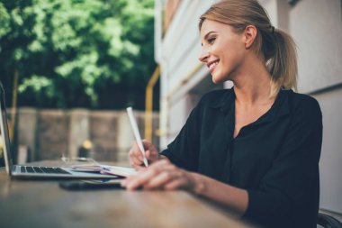Yazı yazarken gülümseyen ve dışarıdaki kafede dizüstü bilgisayarla çalışan günlük tişörtlü sarışın kadın serbest çalışanın yan görüntüsü