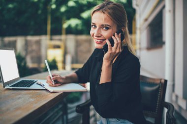Başarılı genç bayan girişimci, günlük giysiler içinde gülümseyip kameraya bakarken cep telefonundan arıyor ve açık kafedeki boş ekran laptopunu kullanıyor. 