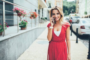 Kırmızı benekli elbiseli, gülümseyen genç bir kadın modern şehrin caddesinde kameraya bakarak akıllı telefondan sohbet ediyor.