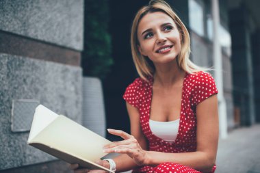 Kırmızı benekli elbiseli, rüya gibi gülümseyen kadın. Kitap okuyor. Modern şehrin sokaklarında oturuyor, gözlerini kaçırıyor.