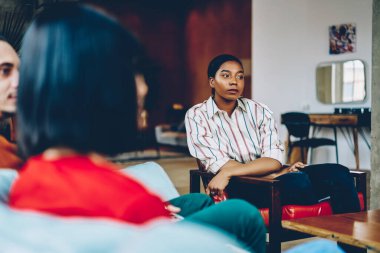 Rahat bir oturma odasında arkadaşlarıyla iletişim kurarken rahat koltukta oturan ve gözlerini kaçıran, sıradan giysili genç Afrikalı Amerikalı kadın.