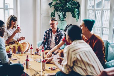 Bir grup çok ırklı insan, arkadaş toplantısı sırasında masada poker oynarken cips koyup alkol içiyorlar.