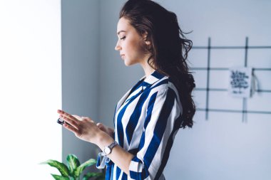 Moda elbiseli şık, uzun kıvırcık saçlı bir kadın blogcunun evde cep telefonuyla internet üzerinden çalışmasının yan görüntüsü.