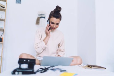 Normal giysiler içinde gülümseyen, telefonla konuşan ve bacak bacak üstüne atarak beyaz duvarda ışık minimalist yatak odasında dizüstü bilgisayarla yazı yazan güzel bir kadın.