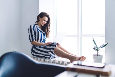 Genç, siyah saçlı, çizgili, modaya uygun elbisesi olan, modern odada büyük pencerenin yanında oturmuş bilgisayardan gülümseyen ve sohbet eden bir kadın.