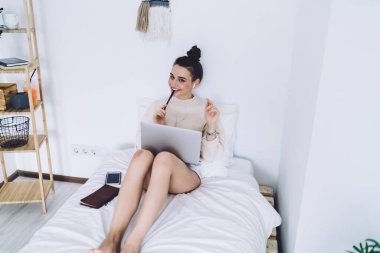 Koyu renk saçlı neşeli bir kadın, beyaz yatağında dizüstü bilgisayarı ve elinde kalemle kameraya bakarken gülümsüyor.