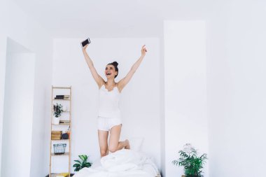 Hevesli bir kadın gece elbisesi içinde rahat bir yatakta el kaldırarak zıplarken aynı zamanda minimalist iç mekanı olan bir odada akıllı telefon kullanıyor ve yukarı bakıyor.