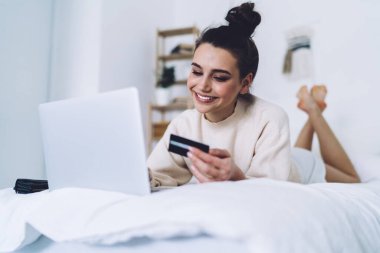 Bej kazaklı, beyaz şortlu, dizüstü bilgisayar kullanırken kredi kartına odaklanan ve internette alışveriş yapan mutlu yetişkin bir kadın. 