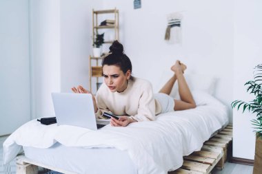 Kafası karışmış, sıradan kıyafetler içinde, kredi kartı tutan ve bilgisayarla online alışveriş yapmaya çalışan bir kadın. Hafif bir dairede, bulanık arka planda yatarken.