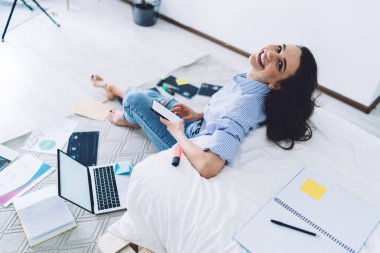 Yukardan gülümseyen, günlük kıyafetleriyle evrak işleriyle uğraşan, akıllı telefonları ve bilgisayarları karıştıran, boş ekranlı, modern bir dairede çıplak ayakla oturan bir kadından.