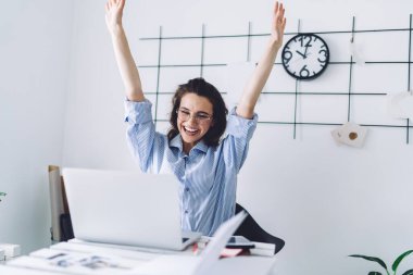 Gözlüklü ve mavi tişörtlü neşeli genç bir kadın ellerini kaldırmış sınıfta gülümsüyor ve asılı saatle dizüstü bilgisayar seyrediyor.