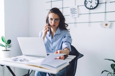 Gözlüklü, mavi elbiseli, cep telefonuyla konuşan ve iş yerindeki not defteri sayfasını okuyan endişeli genç bir kadın.