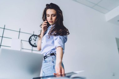 Genç, uzun saçlı, günlük giysiler içinde cep telefonuyla konuşan iş yerinin yakınında duran kağıt parçalarına bakan bir kadından.