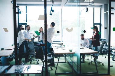 Hedefe ulaşmayı kutlayan bir grup neşeli iş arkadaşına camdan bak ve ofiste cam duvarla otururken mutlu bir şekilde gülümse.
