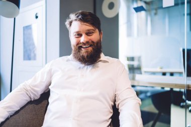 Beyaz gömlekli sakallı yakışıklı adam modern ofiste cam duvara yaslanmış kameraya bakarken gülümsüyor. 