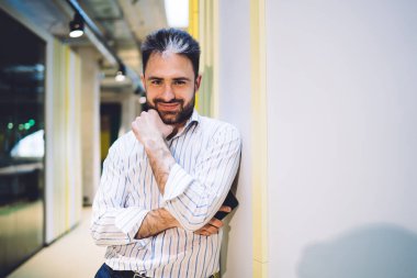 Beyaz gömlekli, memnun genç adam koridorda kameraya bakarken elini sakala koyuyor duvara yaslanıyor. 