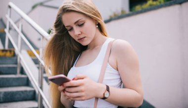 Uzun kolsuz beyaz gömlekli, odaklanmış sarışın kadın yazın merdivenlerde cep telefonuyla mesajlaşıyor.