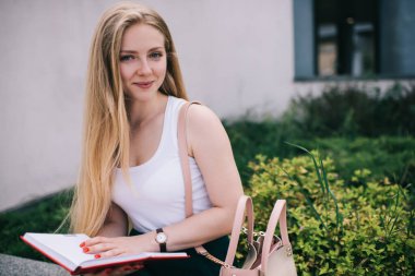 Uzun saçlı, pembe çantalı, kolsuz beyaz gömlekli ve yeşil etekli, mutlu, gülümseyen sarışın kadın yazın kırmızı kapakta kitap okuyor.