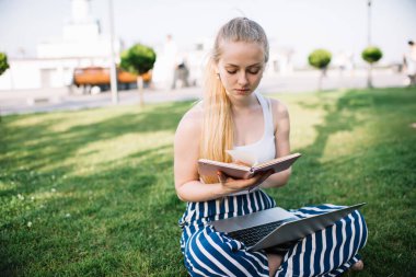 Şehirdeki yeşil çimlerde dizlerinin üstüne çöküp haftanın günlüğünü kontrol eden ciddi konsantre kadın serbest yazar.