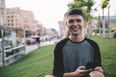 Mutlu, genç, dikkatsiz bir adam kameraya bakıyor ve eğlencenin ve parkta çimlerin üzerinde oturmanın tadını çıkarırken akıllı telefonuyla mesajlaşıyor.