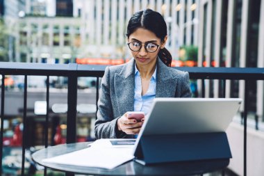 Zarif kıyafetli ve gözlüklü zeki etnik iş kadını New York 'un terasında tablet üzerinde çalışırken açık havada masada oturmuş cep telefonuyla sörf yapıyor. 