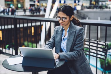 Gri ceketli ve gözlüklü kendine güvenen etnik iş kadını New York 'ta, sokak terasında masada otururken tablet üzerinde çalışıyor.