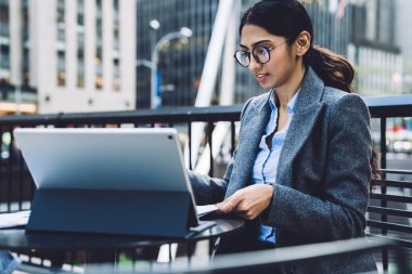 Zarif kıyafetli ve gözlüklü zeki etnik iş kadını New York 'un sokak terasında tableti incelerken açık havada masalarda oturup belgeleri kontrol ediyor. 