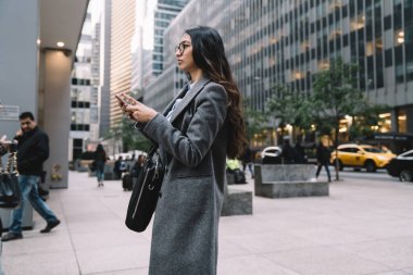 New York caddesinde yürüyen ve cep telefonu kullanan gözlüklü genç bir iş kadınının yan görüntüsü.