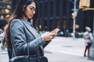 New York şehrinin arka planında dikilirken, uzun saçlı, modaya uygun ceketli, sörf yapan genç bayanın yan görüntüsü. 