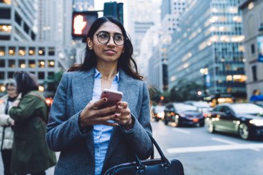 Yuvarlak gözlüklü, zarif, genç, esmer kadın. Siyah çanta ve gri ceket. New York 'ta yaya geçidinin yanındaki akıllı telefona dokunuyor ve başka yöne bakıyor.