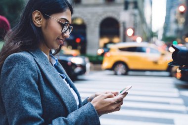 Yuvarlak gözlüklü, gri montlu, New York 'un kalabalık caddesinde, bulanık arka planda yaya geçidinde akıllı telefondan konuşan esmer bir kadının yan görüntüsü.