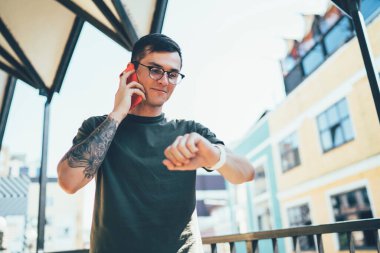 Gözlüklü, haki rengi tişörtlü, kol saatlerini kontrol eden ve balkonda cep telefonuyla konuşan sıska, zeki bir erkek.