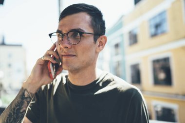 İşbirlikçi proje ile tartışmak için akıllı telefonu kullanarak dövmeli, gündelik giysiler ve gözlüklü, havalı bir erkek.