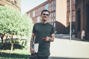 Gözlüklü ve elinde dövmesi olan, dizüstü bilgisayarını elinde tutarken ve sokak ortasında dururken kameraya bakıp cep telefonu kullanan kendinden emin genç, serbest çalışan bir erkek.