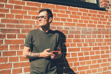 Gözlüklü genç erkek öğrenci akıllı telefon kullanırken ve gün ışığında kırmızı tuğladan duvara yaslanırken başka yöne bakıyor.