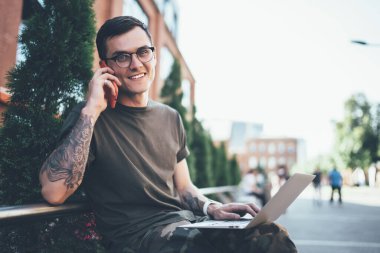 Genç, gözlüklü, kollarında dövmeleri olan dizlerinde dizleri olan, cep telefonuyla konuşan ve binanın yanındaki bankta oturan neşeli bir adam.