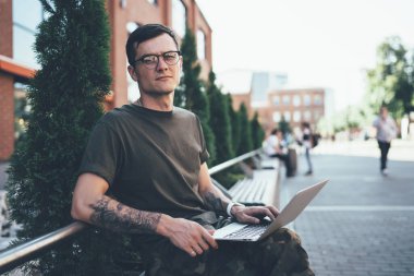 Gözlüklü, dövmeli, kameraya bakan ve dar sokaktaki bankta otururken dizüstü bilgisayar kullanan kendine güvenen genç adam.