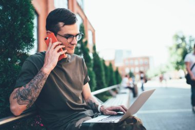 Park sokağındaki bankta otururken cep telefonuyla konuşan ve dizüstü bilgisayar kullanan odaklanmış genç bir erkek öğrenci.