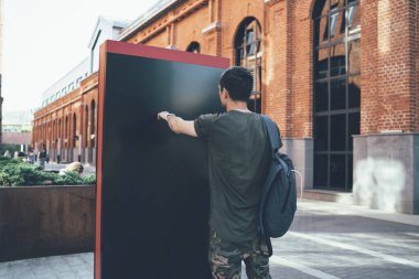 Gündelik giysiler içinde, sırt çantasıyla sokak ortasında duran boş reklam panosuna bakan genç bir erkek.
