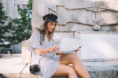 Konsantre olmuş, açık beyaz elbise giyen, elinde kamera olan genç bir bayan yaz günü caddede, anıtın yanında harita kontrol ediyor.