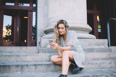 Gülümseyen, uzun saçlı, cep telefonuyla mesaj atan genç bir bayan yaz günü merdivenlerde oturuyor. 