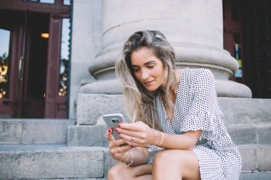 Gülümseyen, uzun saçlı, cep telefonuyla mesajlaşan genç bayan yaz gününde granit merdivenlerde oturuyor. 