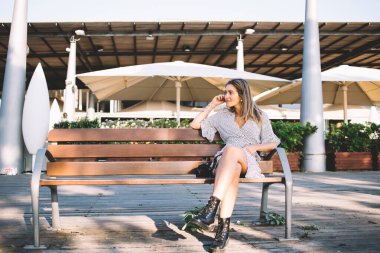 Günlük kıyafetleri ve siyah ayakkabıları olan pozitif bir kadın arkadaşlarıyla bacak bacak bacak üstüne atarak yazlık kafe hakkında bankta oturuyor.