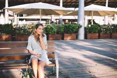 Şık bir yaz elbisesi içinde gülümseyen, neşeli bir kadın ahşap bir bankta otururken saçlarına dokunuyor ve cep telefonuyla diğer tarafa bakıyor.