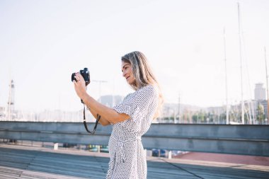 Uzun saçlı, açık renk elbiseli, puantiyeli, siyah kameralı gülümseyen bir kadın yazın, arka planda şehirle birlikte tahta zeminde fotoğraf çekiyor.