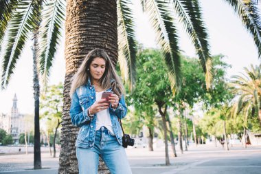 Kotlu genç bayan kamerayla takıyor. Güneşli bir günde arkadaşlarıyla parkta yürürken mesajlaşmak için cep telefonu kullanıyor.