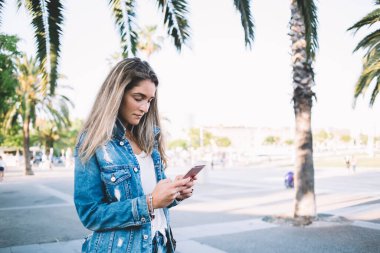 Kot pantolon giymiş genç bir kadın güneşli bir günde parkta yürürken arkadaşlarıyla mesajlaşmak için cep telefonu kullanıyor.