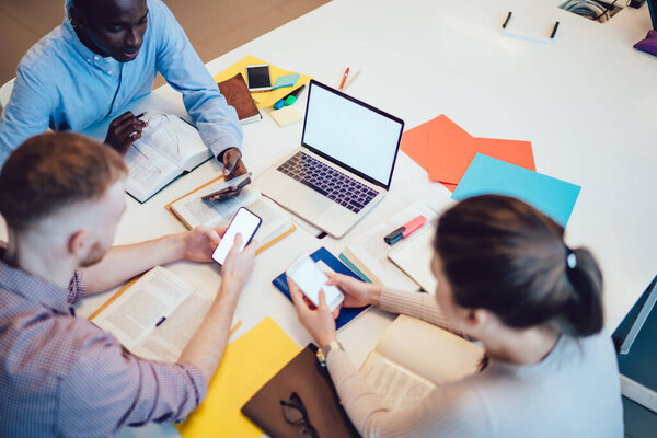 From above serious diverse coworkers in casual wear sitting at desk in office and browsing smartphone while surfing laptop and reading book